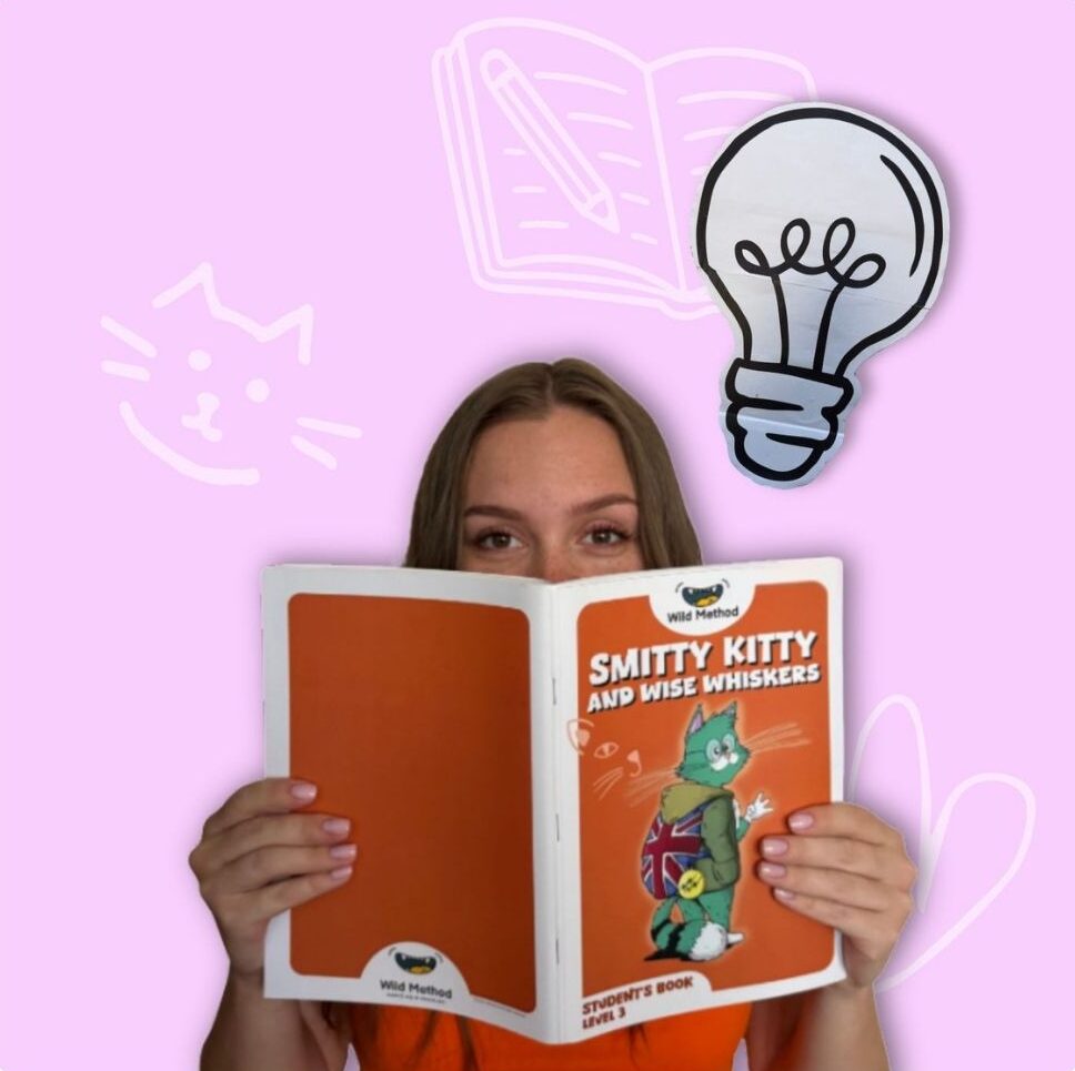 Dziewczyna w pomarańczowej koszulce trzymająca podręcznik „Smitty Kitty and Wise Whiskers” wydawnictwa Wild Method. Nad jej głową unosi się komiksowa żarówka.