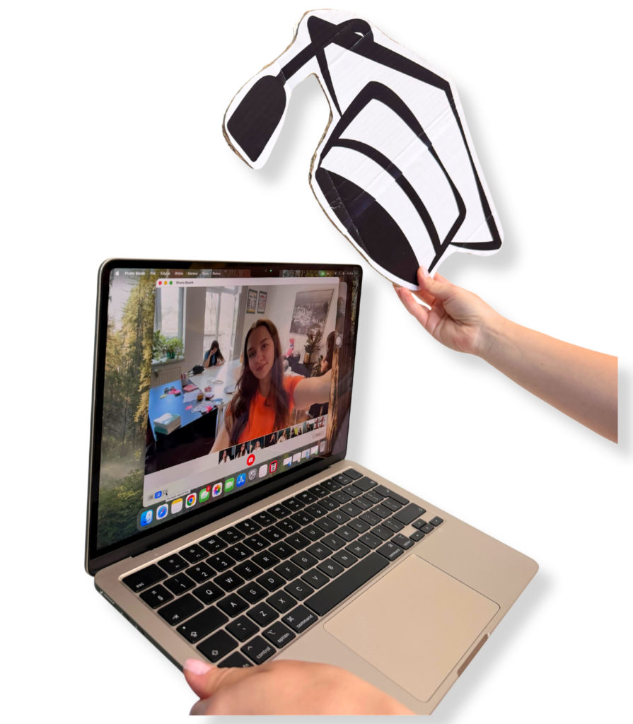Widok z perspektywy ekranu laptopa na dziewczynę prowadzącą zajęcia online. Nad laptopem ręka trzyma komiksowy biret absolwenta.